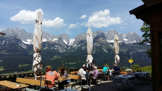 Going_Hollenauer Kreuz_Terasse_Sommer_2022_5 Eine schöne Berglandschaft mit schneebedeckten Gipfeln und einem klaren Himmel. Auf einer Terrasse sitzen Gäste und genießen die Aussicht.