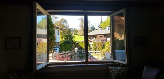 Blick aus dem Wohnzimmer Blick durch ein geöffnetes Fenster auf benachbarte Häuser, Gärten und eine grüne Wiese unter blauem Himmel.