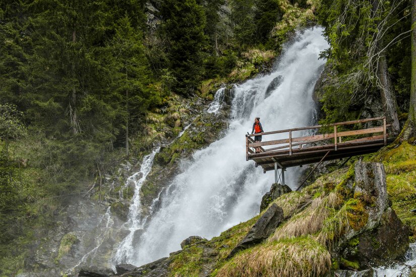 Sintersbach Ein beeindruckender Wasserfall umgeben von grünen Bäumen. Eine Person steht auf einer Holzplattform und genießt die Aussicht.