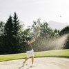 Ein Golfer schlägt mit Schwung aus einem Sandbunker auf dem Golfplatz Kitzbühel-Schwarzsee-Reith, während Sand in die Luft wirbelt und die Sonne scheint.