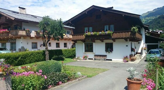 Reason Sommer Außenansicht Zweistöckiges Alpenhaus mit weißer Fassade, Holzbalkon und Blumenschmuck vor einer Bergkulisse.