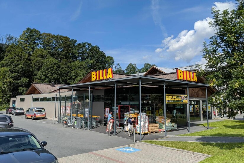 Außenansicht eines Billa-Supermarktes mit Parkplatz und bewaldetem Hintergrund unter blauem Himmel.