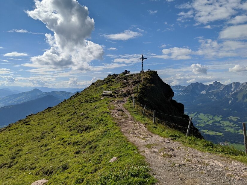 Horn Gipfel-Höhenweg Schmaler Wanderweg führt über einen grasbewachsenen Grat zu einem Gipfelkreuz unter blauem Himmel.