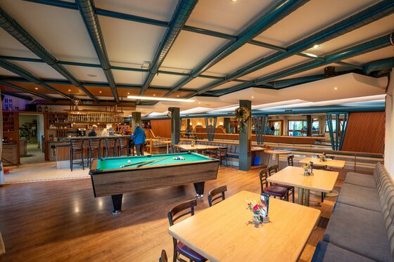 Innenansicht eines Gastraums mit Billardtisch, Holztischen und einer Bar im Hintergrund.
