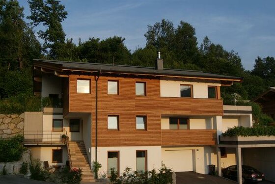 Unterkunft Genner im Sommer Modernes mehrstöckiges Gebäude mit Holzfassade und weißem Putz vor bewaldetem Hintergrund.