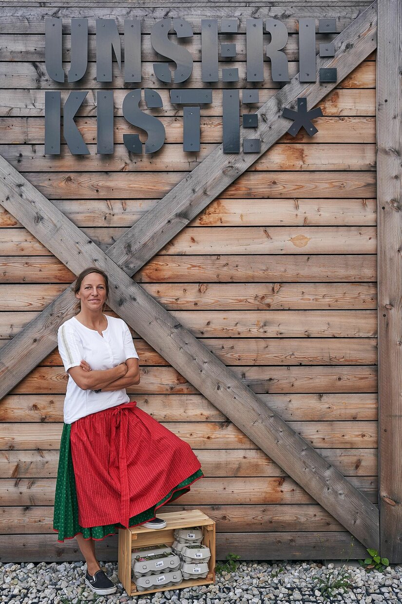 Unsere Kiste Eine Frau in Tracht steht vor einer Holzwand mit dem Schriftzug Unsere Kiste.