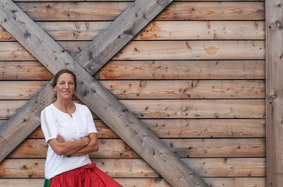 Eine Frau in Tracht steht vor einer Holzwand mit dem Schriftzug Unsere Kiste.