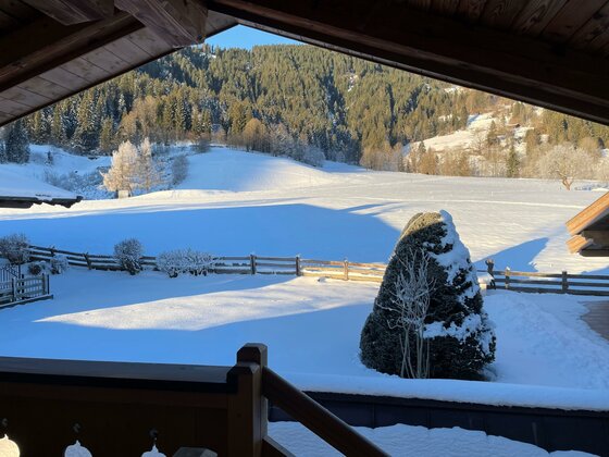 Aussicht Loipe Kaiser Loft Blick von einem Holzbalkon auf eine verschneite Wiese, einen Zaun und bewaldete Berge im Hintergrund.