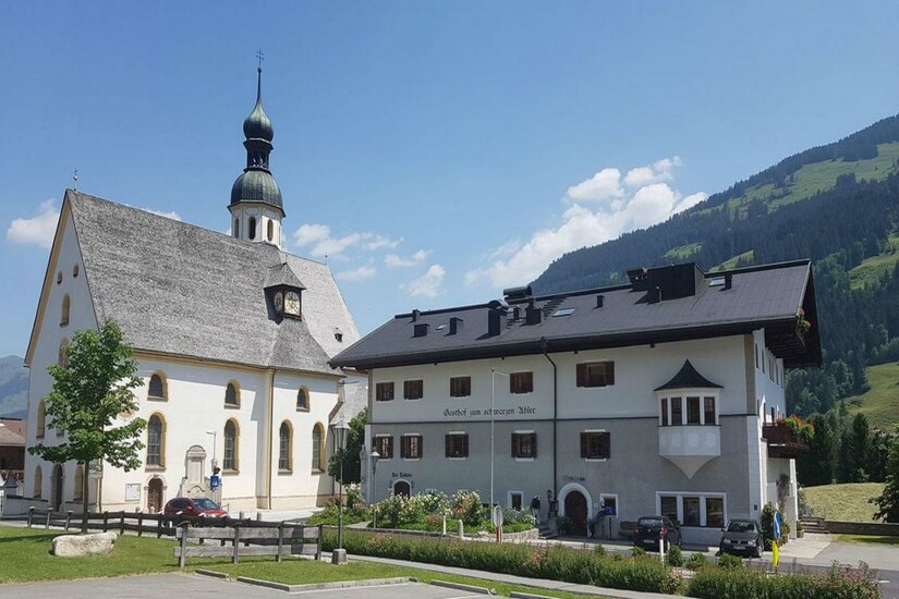 Eine malerische Kirche mit einem hohen Turm steht neben einem traditionellen Gebäude. Im Hintergrund sind sanfte Hügel und ein klarer Himmel zu sehen.