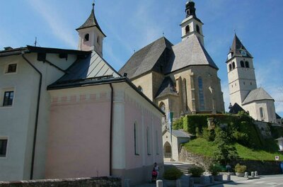 Spitalskirche Ein historisches Gebäude mit mehreren Türmen und unterschiedlichen Dachformen. Im Vordergrund ist ein helles Haus zu sehen, das sich an eine grüne Hügelkuppe anschmiegt.
