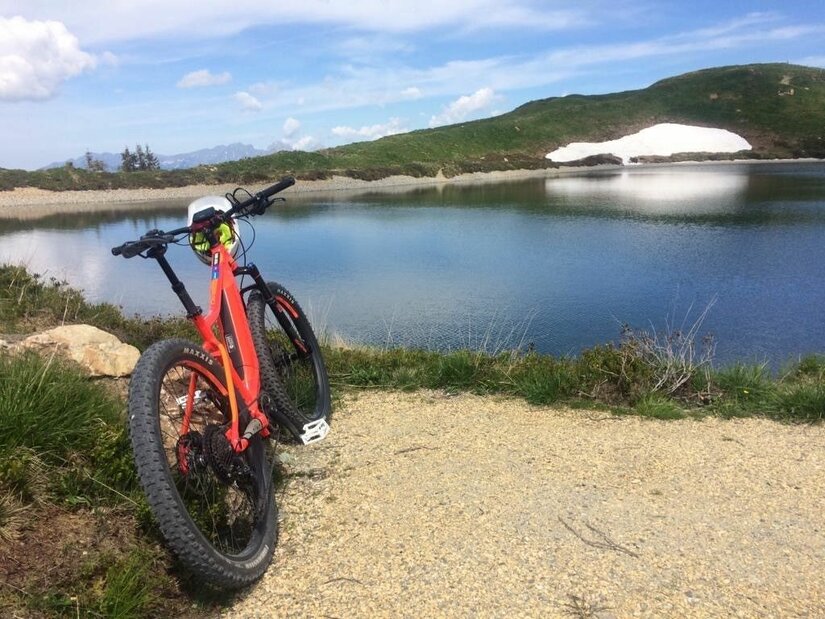 Mountainbike am Saukaser-Pengelstein Ein orangefarbenes Mountainbike steht auf einem Schotterweg vor einem Bergsee mit Schneerest am Ufer.