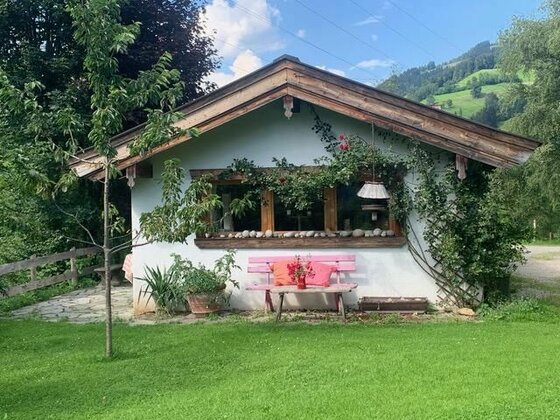 Kleines weißes Gartenhaus mit Holzdach, rosa Sitzbank und Blumen vor einer grünen Berglandschaft.