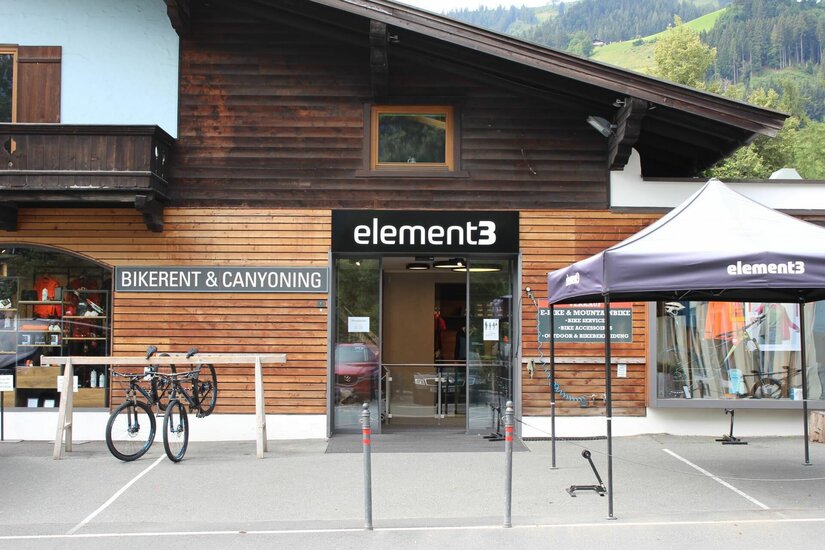 Eingangsbereich eines Sportgeschäfts mit Holzfassade, Fahrrädern im Ständer und einem schwarzen Zeltdach.