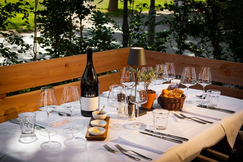 Chizzo Terrasse Gedeckter Tisch im Freien mit Weingläsern, einer Weinflasche, Besteck und Brotkorb vor einer Holzbank.