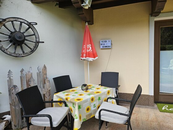 Außenterrasse mit Tisch, vier Stühlen, Sonnenblumendecke und einem dekorativen Wagenrad an der Wand.