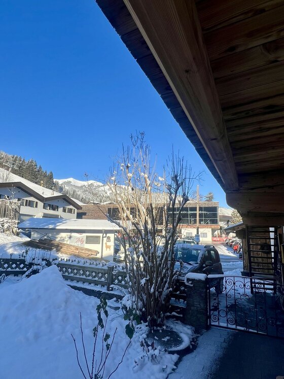 Blick von einem Holzhaus auf eine verschneite Siedlung mit Bergen im Hintergrund unter blauem Himmel.