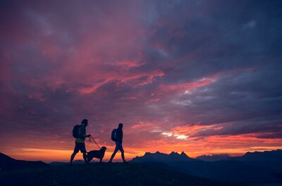Zwei Wanderer und ein Hund spazieren bei Sonnenuntergang in den Bergen. Der Himmel ist in wunderschönen Farben gehalten.