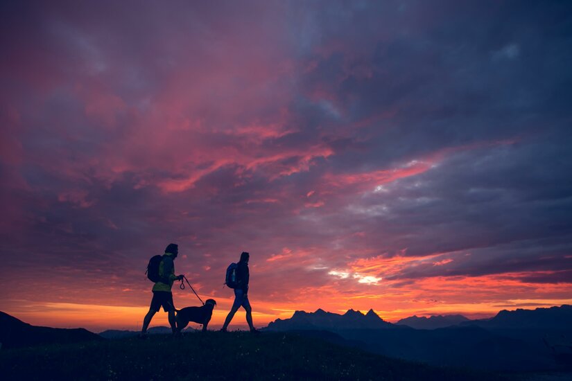 Zwei Wanderer und ein Hund spazieren bei Sonnenuntergang in den Bergen. Der Himmel ist in wunderschönen Farben gehalten.
