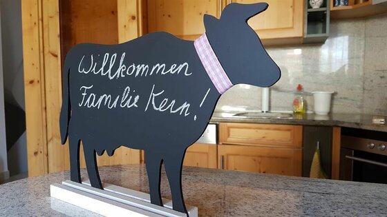 Schwarze Kreidetafel in Form einer Kuh mit Willkommensgruß auf einer Arbeitsplatte in einer Küche.