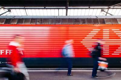 Bahnhof Kitzbühel-Schwarzsee Seitenansicht eines roten Zuges am Bahnsteig mit verschwommenen Personen im Vordergrund.