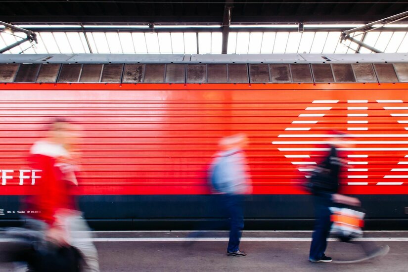 Bahnhof Kitzbühel-Schwarzsee Seitenansicht eines roten Zuges am Bahnsteig mit verschwommenen Personen im Vordergrund.