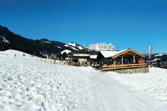 Restaurant Reither Streif im Winter Holzhütte mit Terrasse an einer verschneiten Skipiste vor Bergkulisse.