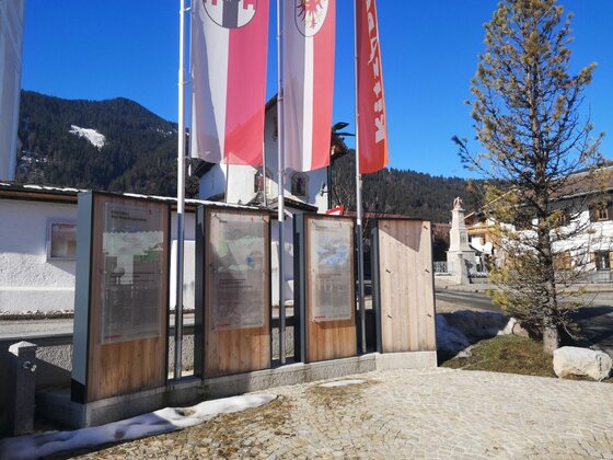 Infopoint Griesbach Infotafeln aus Holz und Glas vor rot-weißen Flaggen im Freien bei Sonnenschein.