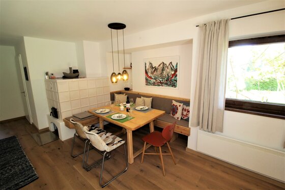 Moderner Essbereich mit Holztisch, Eckbank, Kachelofen und Fenster.