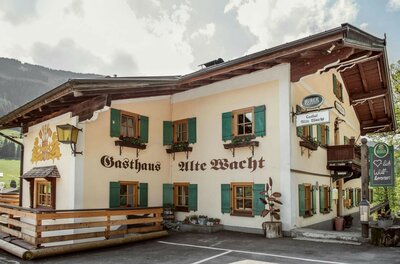Gasthof Alte Wacht im Sommer Zweistöckiges Gasthaus Alte Wacht mit hellen Wänden, grünen Fensterläden und Holzbalkon vor bewaldetem Berg.