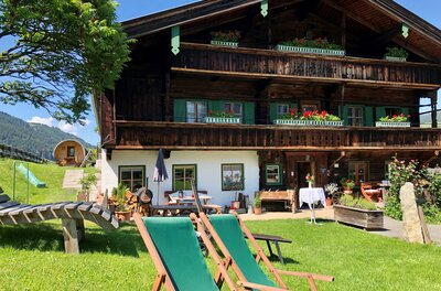 Ein traditionelles Holzhaus umgeben von grüner Wiese und Bäumen. Im Vordergrund stehen bequeme Liegen zum Entspannen.