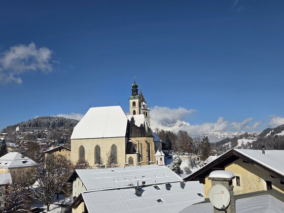 Eine schöne winterliche Landschaft mit einer Kirche und schneebedeckten Dächern. Im Hintergrund sind die Berge und ein blauer Himmel zu sehen.