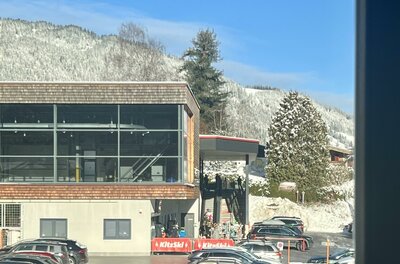 IMG_4610 Ein modernes Gebäude mit großen Fenstern, umgeben von schneebedeckten Bergen. Im Vordergrund sind viele parkende Autos zu sehen.