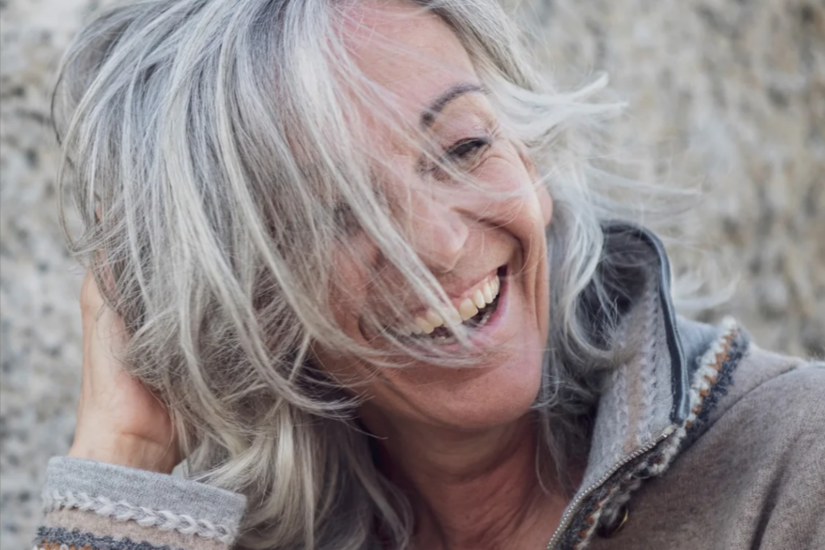 Porträt Sabine Sommeregger Nahaufnahme einer lachenden Frau mit grauen Haaren vor einer Steinwand.