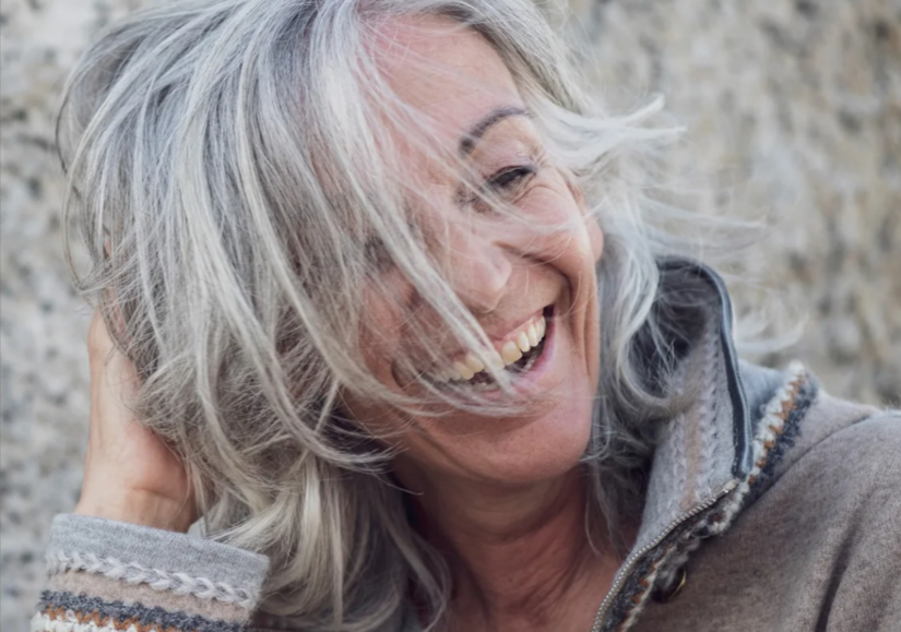 Nahaufnahme einer lachenden Frau mit grauen Haaren vor einer Steinwand.
