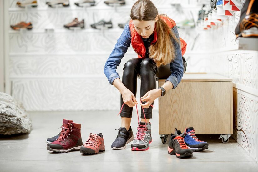 Eine Frau sitzt in einem Sportgeschäft und schnürt einen grauen Wanderschuh mit pinken Akzenten.