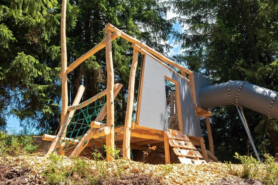 Ein Spielgerüst aus Holz mit Kletternetz, Leiter und einer Röhrenrutsche vor einem Nadelwald.