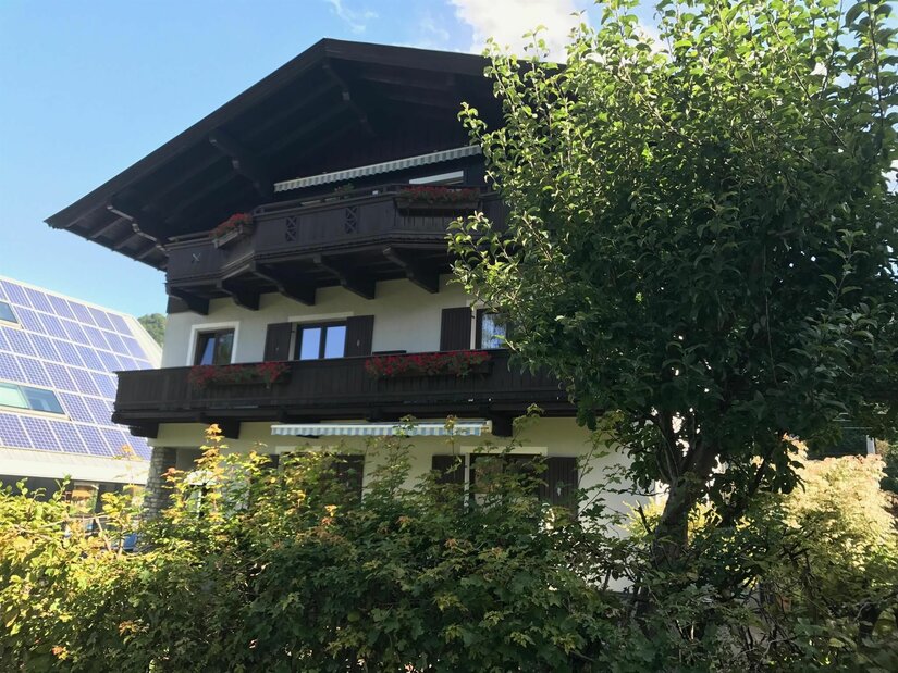 Mehrstöckiges Alpenhaus mit dunklen Holzbalkonen, Blumenkästen und Photovoltaikanlage im Hintergrund.