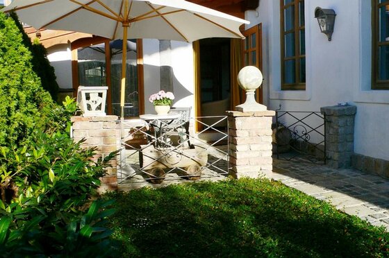 Terrasse Apartment Alpenland Gepflasterte Terrasse mit weißem Sonnenschirm, kleinem Tisch, Blumen und gemauerten Pfosten vor einem Haus.