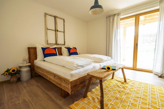 Schlafzimmer Ferienwohnung 3 Schlafzimmer mit Doppelbett aus Altholz, gelbem Teppich und Balkonzugang.