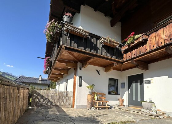 Eingangsbereich eines alpinen Hauses mit Holzbalkon, Blumenkästen und einer kleinen Holzbank auf einer Steinterrasse.