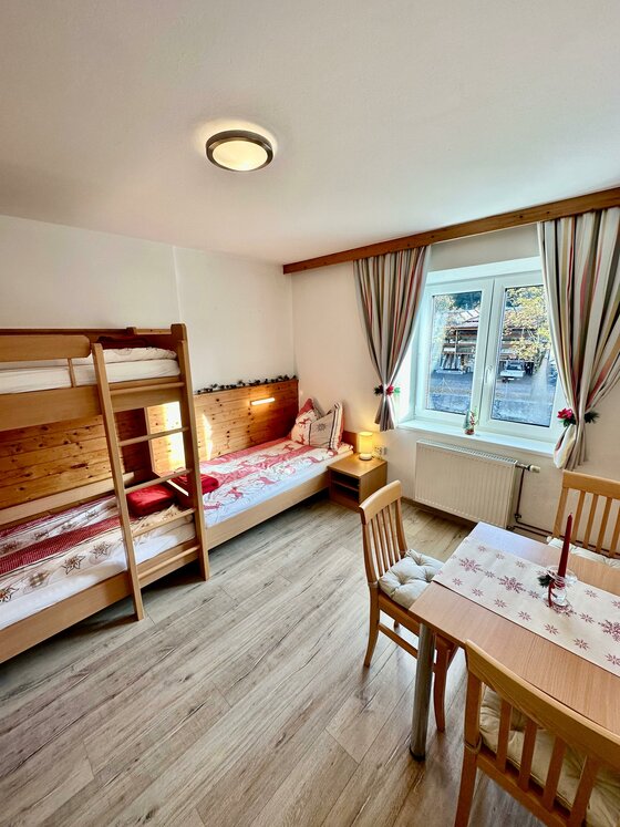 Helles Zimmer mit Holz-Etagenbett, Einzelbett, Esstisch und Fenster.
