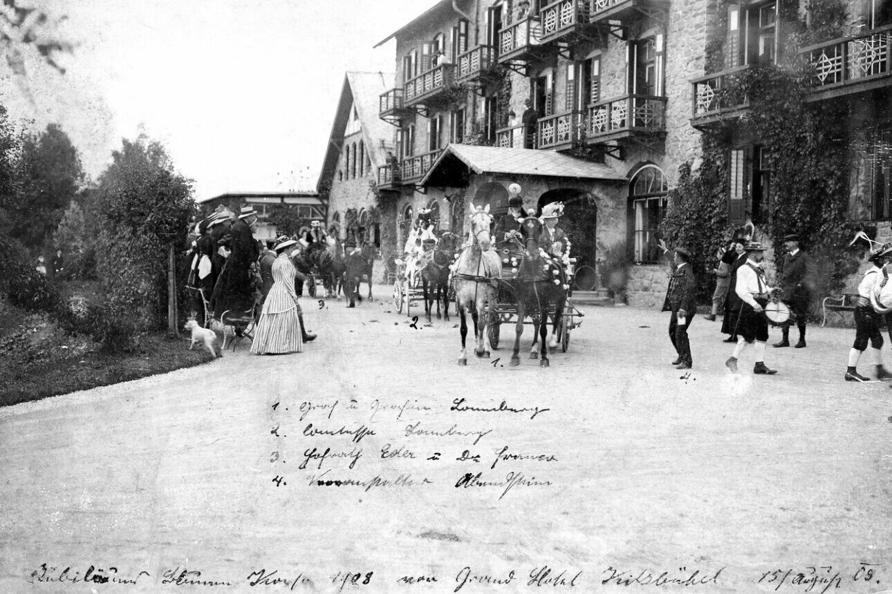 Eine Kutsche mit zwei Pferden und mehrere Menschen stehen vor dem Grand Hotel in Kitzbühel