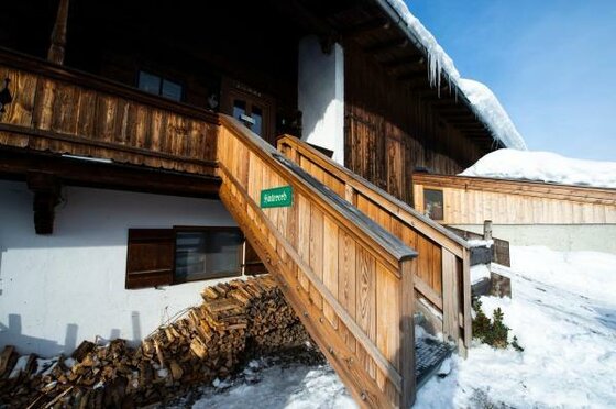 Hölzerner Treppenaufgang zu einem verschneiten Bauernhaus mit Brennholzstapel.