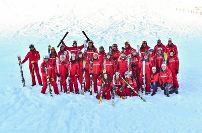 Skischule Jochberg Team Eine Gruppe von Skilehrern in roten Anzügen posiert mit Skiern auf einer verschneiten Piste.