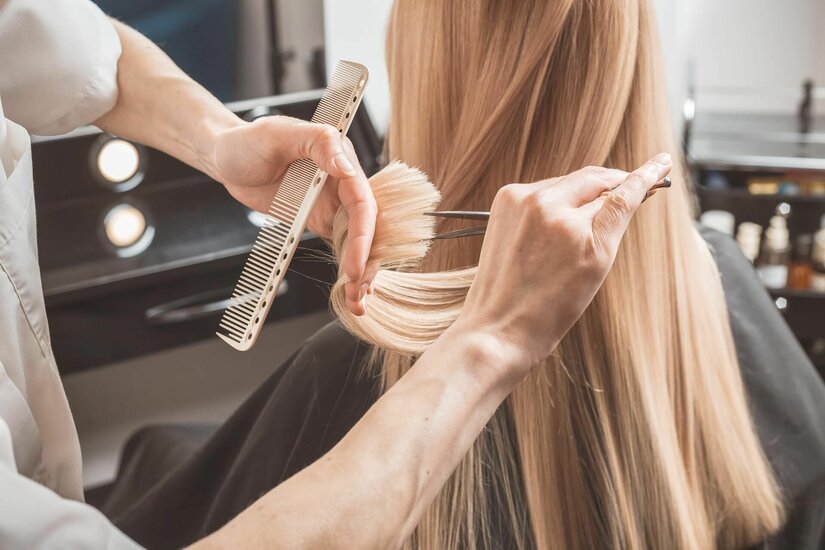 Haarschnitt im Salon Haargenau Nahaufnahme einer Person, die mit Kamm und Schere lange blonde Haare schneidet.
