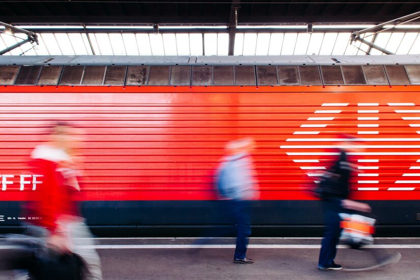 Ein Bahnhof mit einer roten Wand im Hintergrund. Menschen gehen schnell vorbei, während sie unterwegs sind.