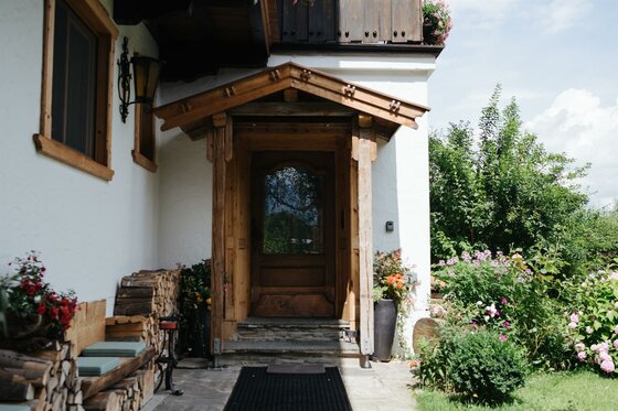 Hauseingang aus Holz mit Vordach, daneben gestapeltes Brennholz und Blumenbeete.