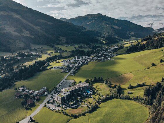 Kempinski Hotel Das Tirol Luftaufnahme eines Hotels in einem grünen Alpental mit angrenzendem Dorf und bewaldeten Bergen.