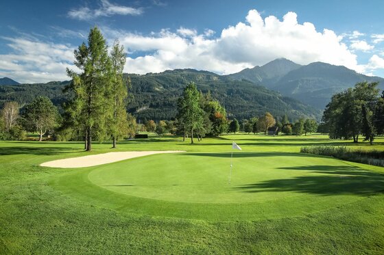Gepflegtes Green auf einem Golfplatz mit Sandbunker vor bewaldeten Bergen und blauem Himmel.