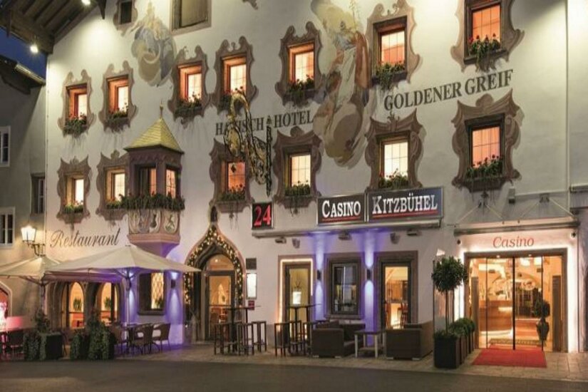 Spätgotisches Portal Ein traditionelles Hotelgebäude mit bunten Fenstern und schöner Fassade. Im Vordergrund ist ein Casino und Außensitzplätze sichtbar.